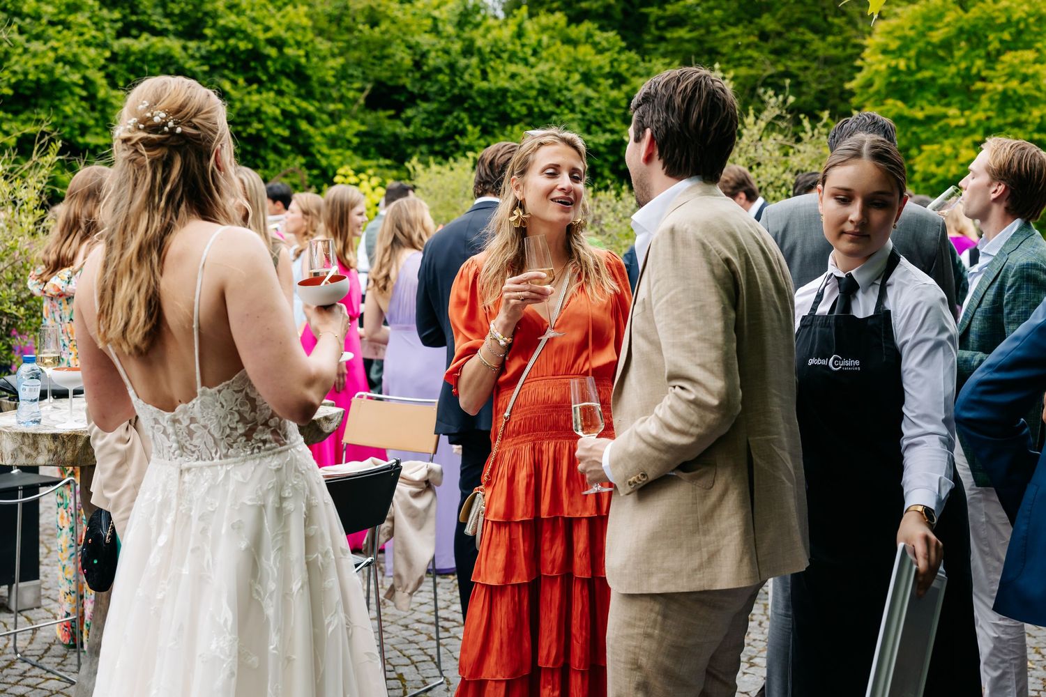 Wedding guests mingle outdoors, including a bride in a white gown and a woman in an orange dress.