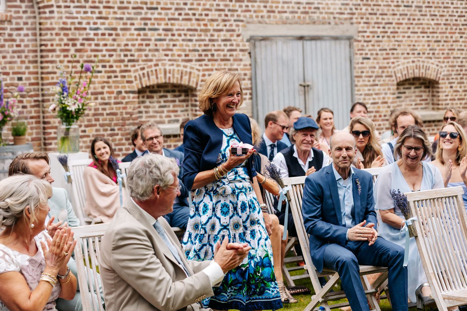 Wedding guest in blue floral dress standing and speaking during outdoor ceremony at brick venue.