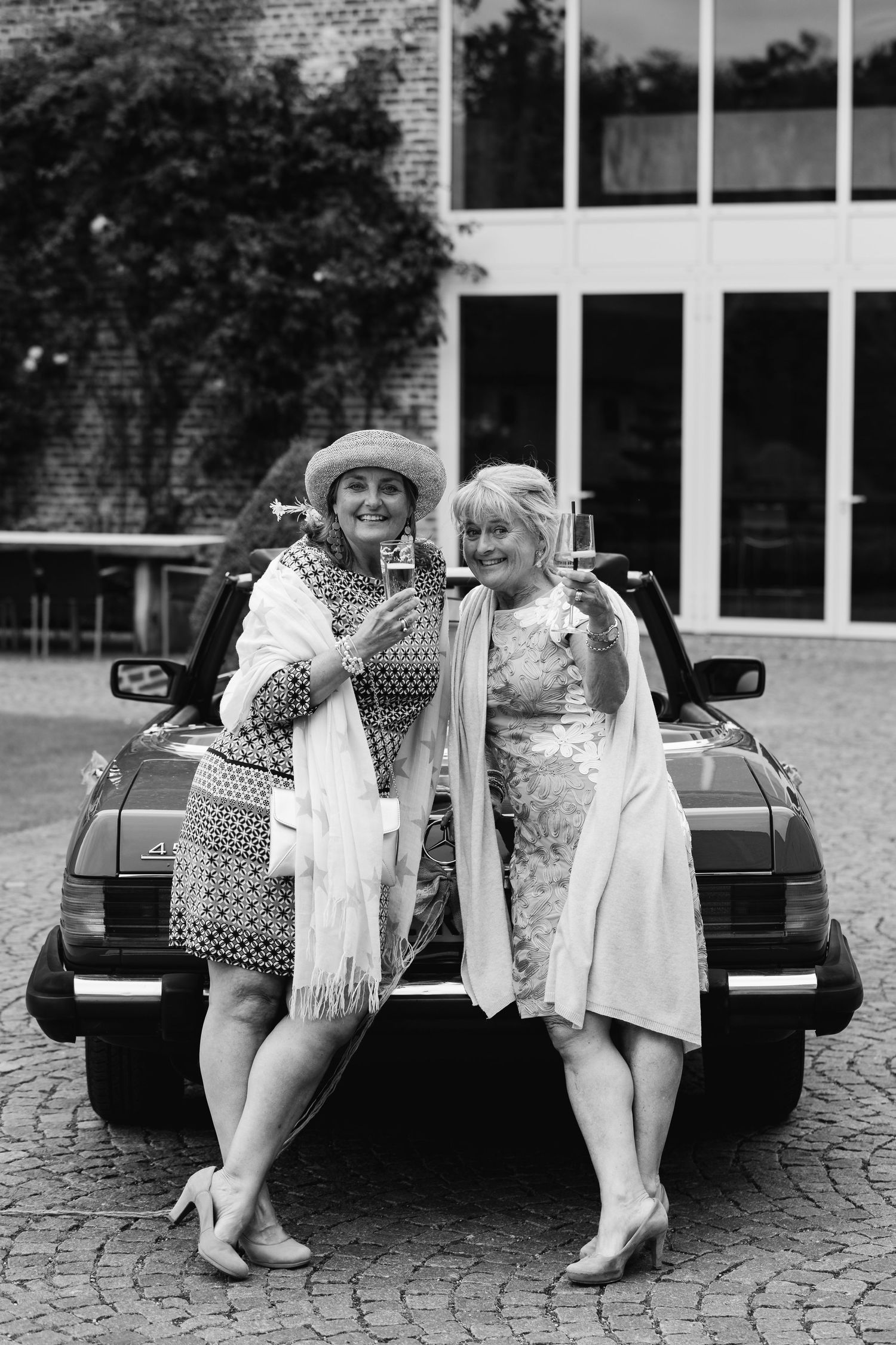 Two older women in elegant dresses posing together leaning against a vintage convertible car.