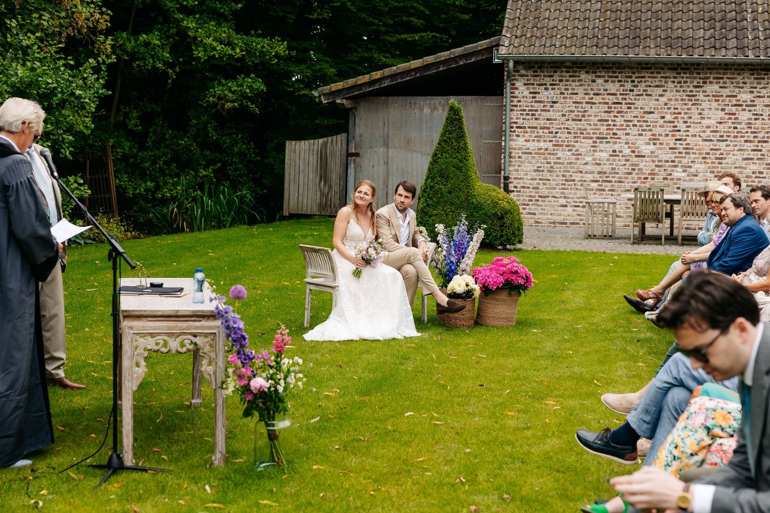 A bride and groom sit together during an intimate outdoor wedding ceremony on a lush green lawn.