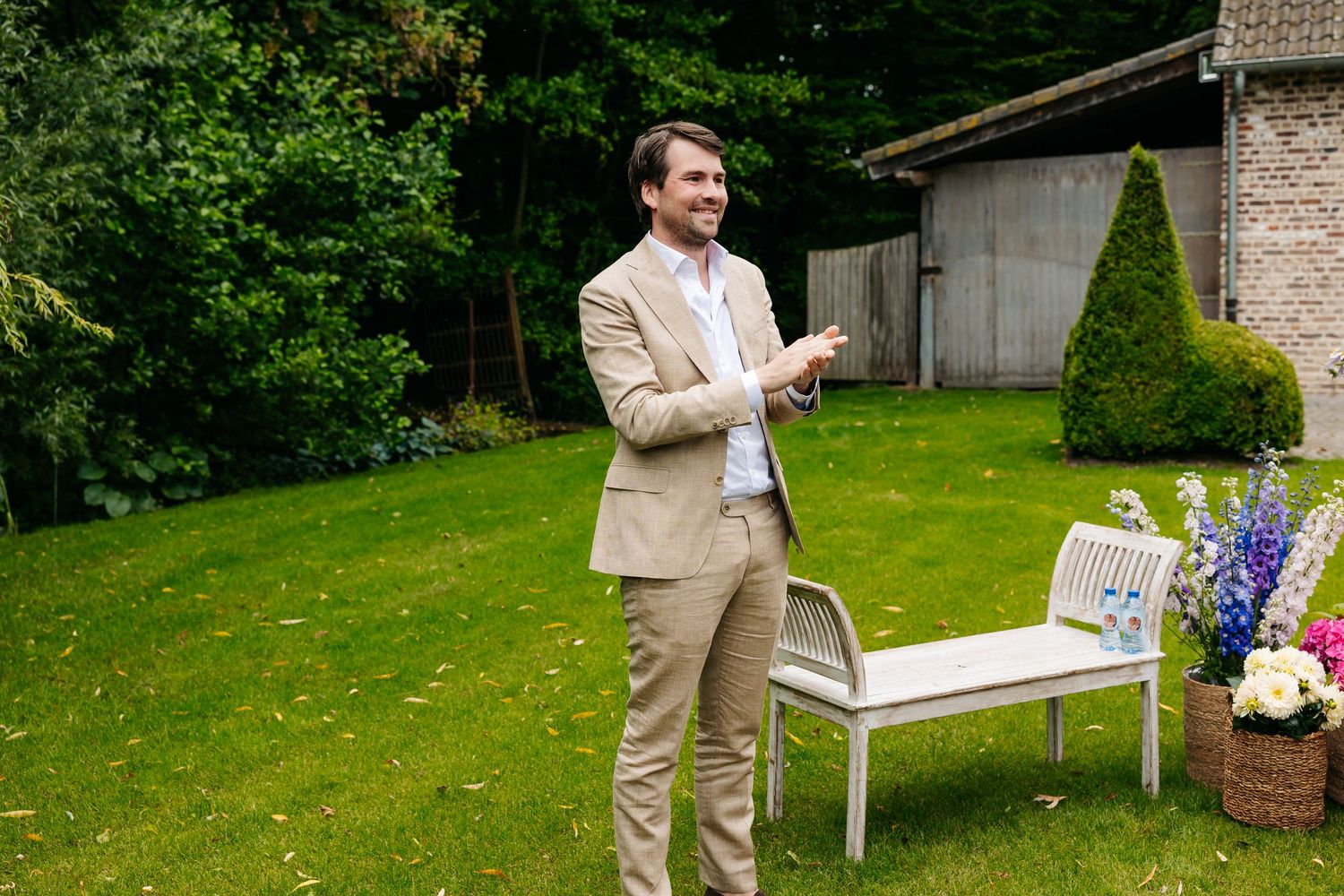 Groom in beige suit stands smiling on green lawn during outdoor wedding ceremony.