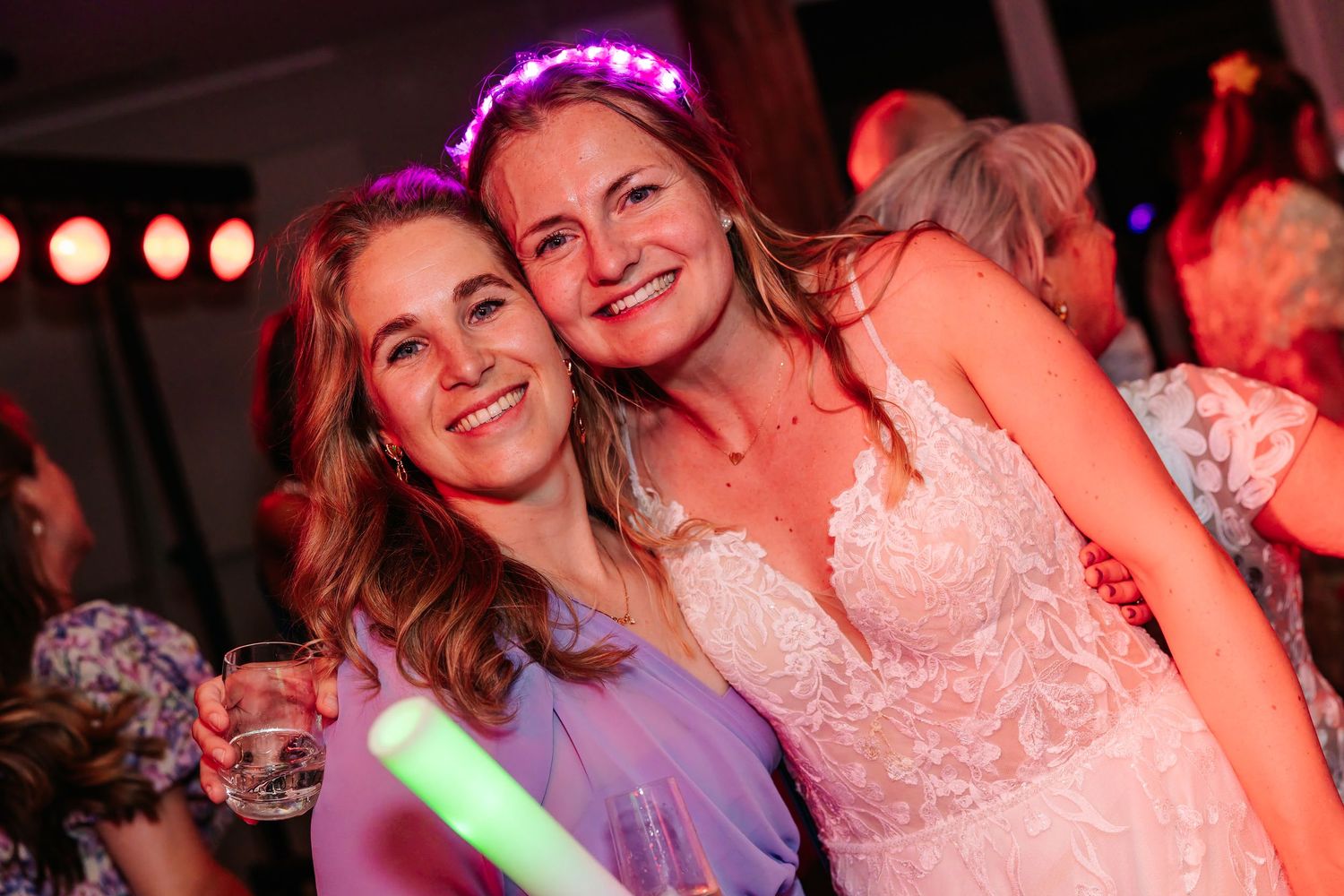 Bride wearing light-up headband poses with smiling friend at colorful evening wedding party.