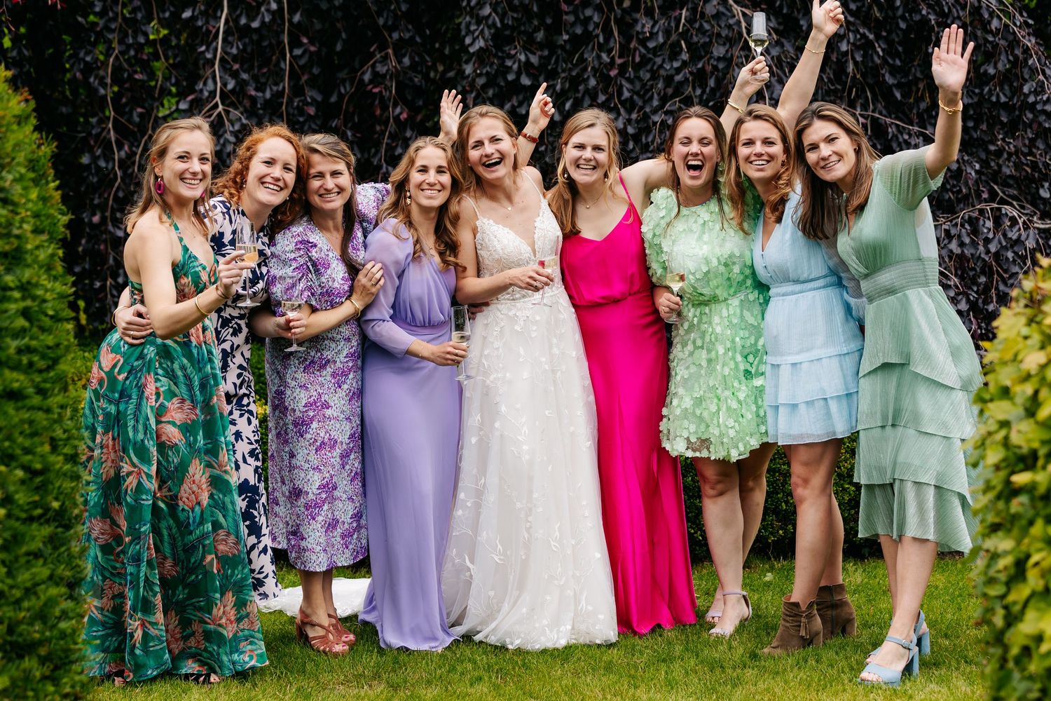 Bride celebrating with bridesmaids in colorful dresses, laughing and raising glasses outdoors.