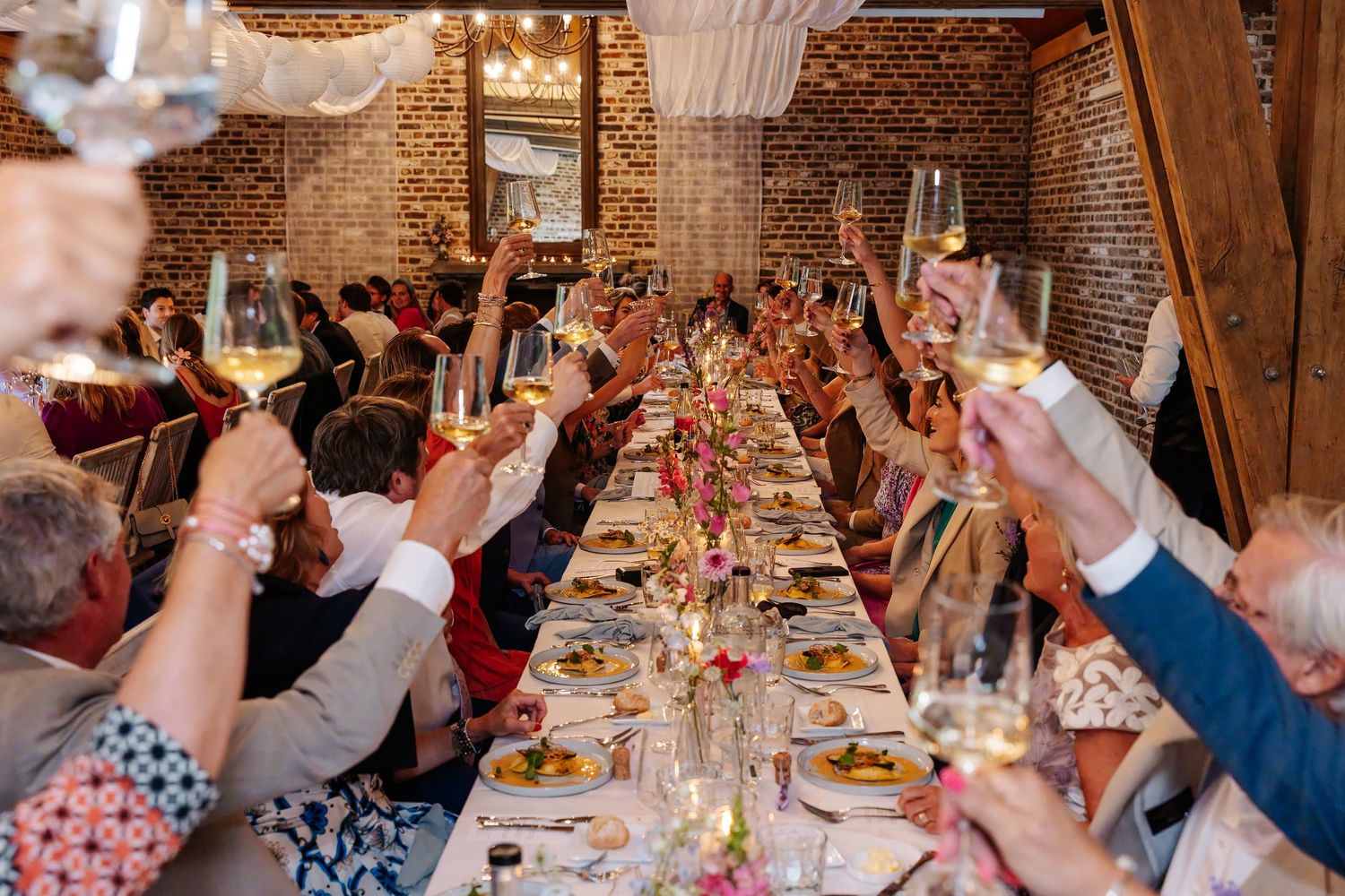 Wedding guests raise champagne glasses in celebratory toast at long decorated dinner table indoors.