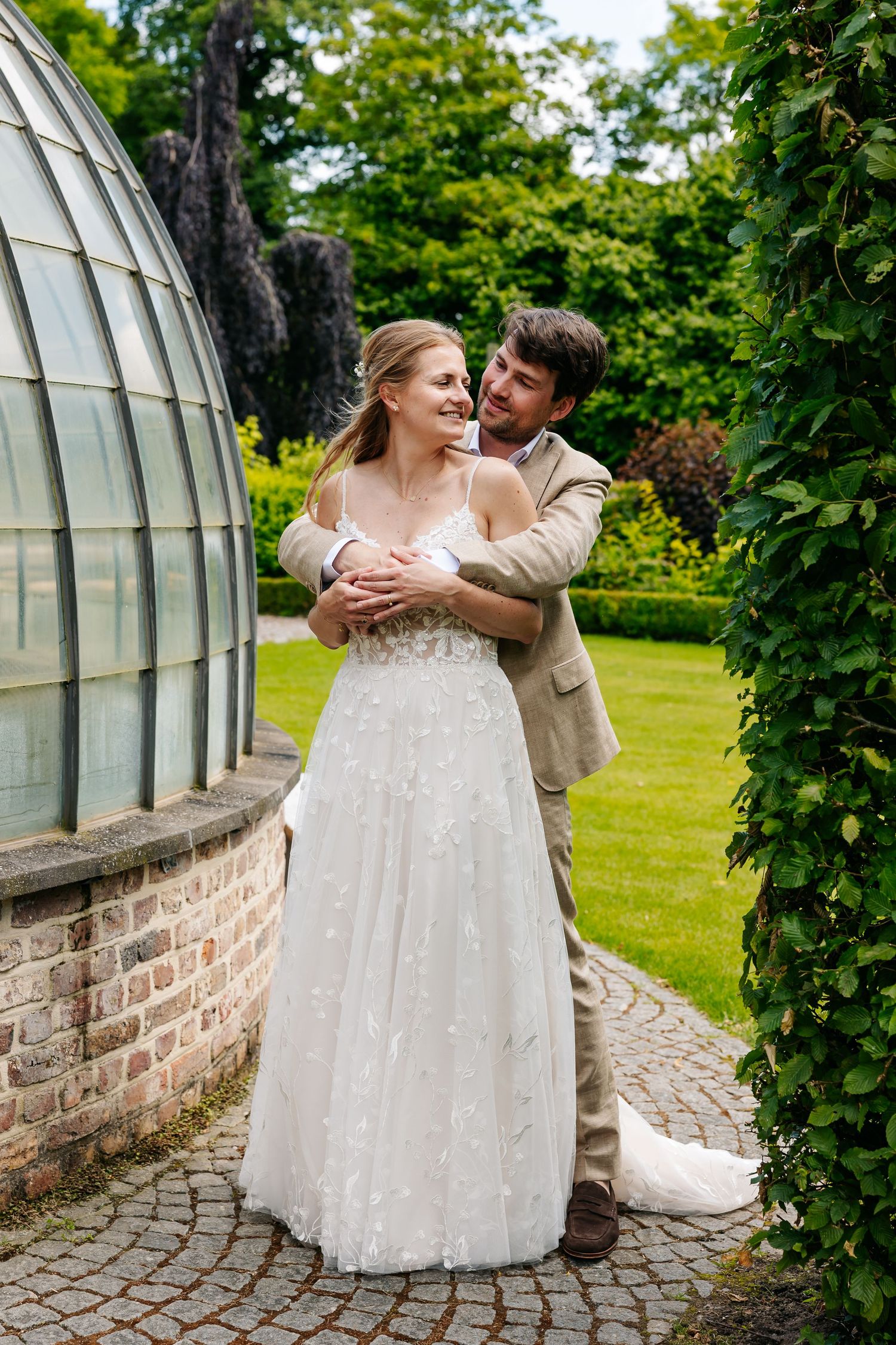Groom embracing bride in floral wedding gown beside glass greenhouse in lush garden.