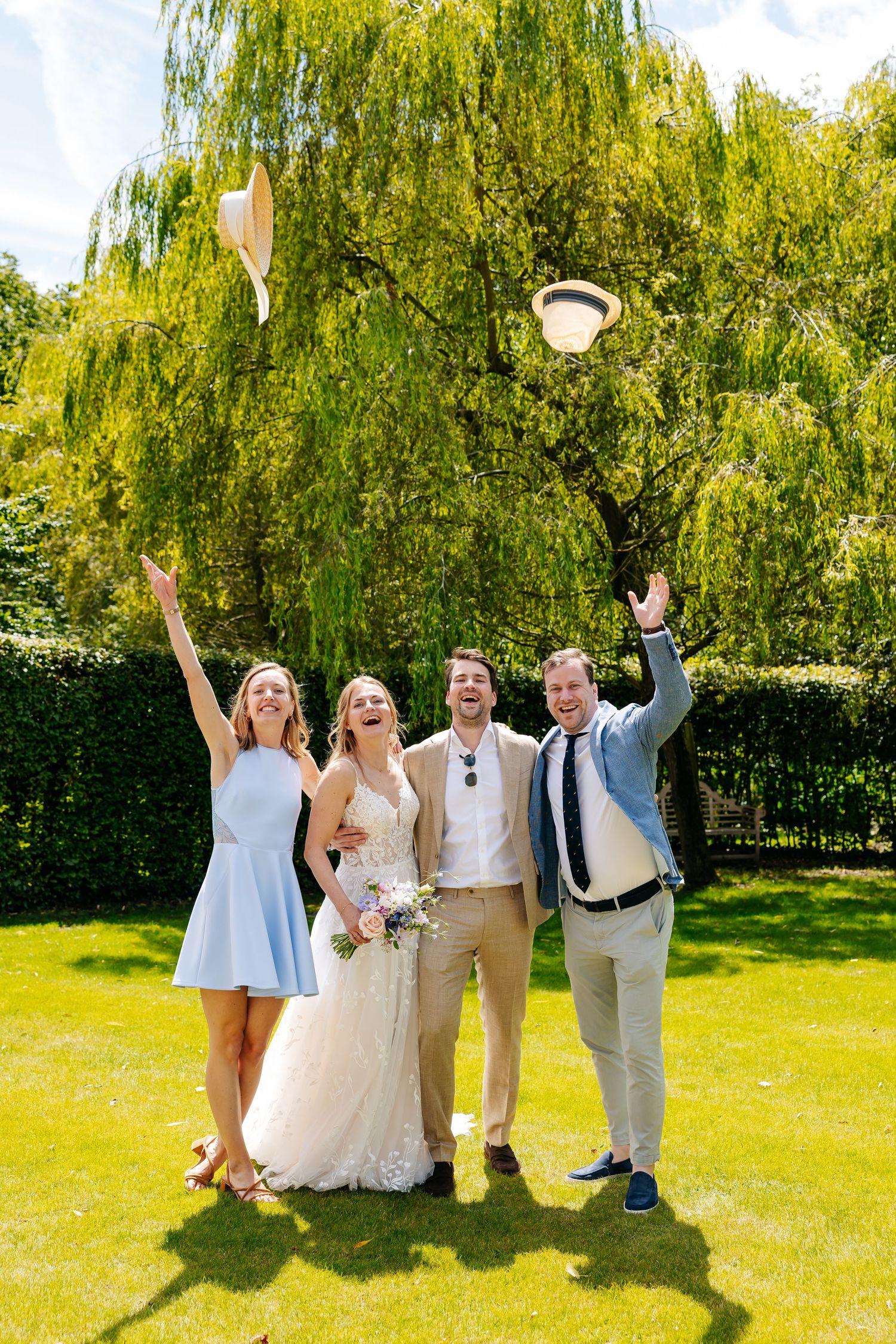 Bride, groom, and guests joyfully tossing hats in the air in sunny garden setting.