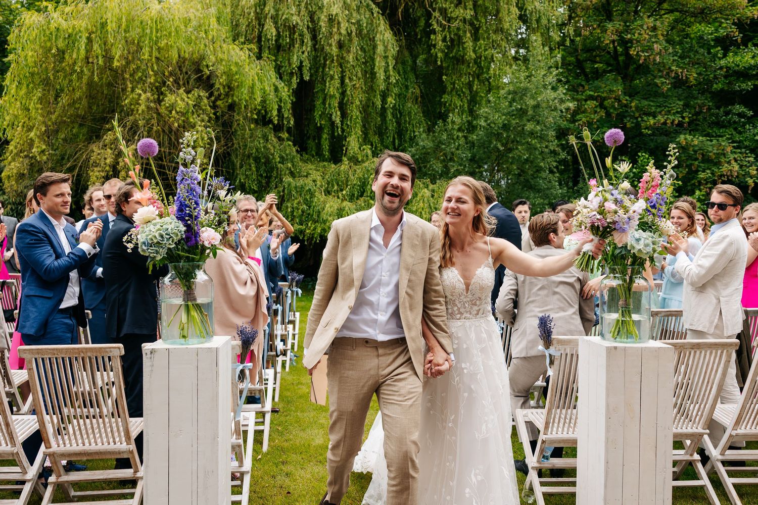 Smiling newlyweds hold hands walking down a flower-lined aisle after their ceremony.