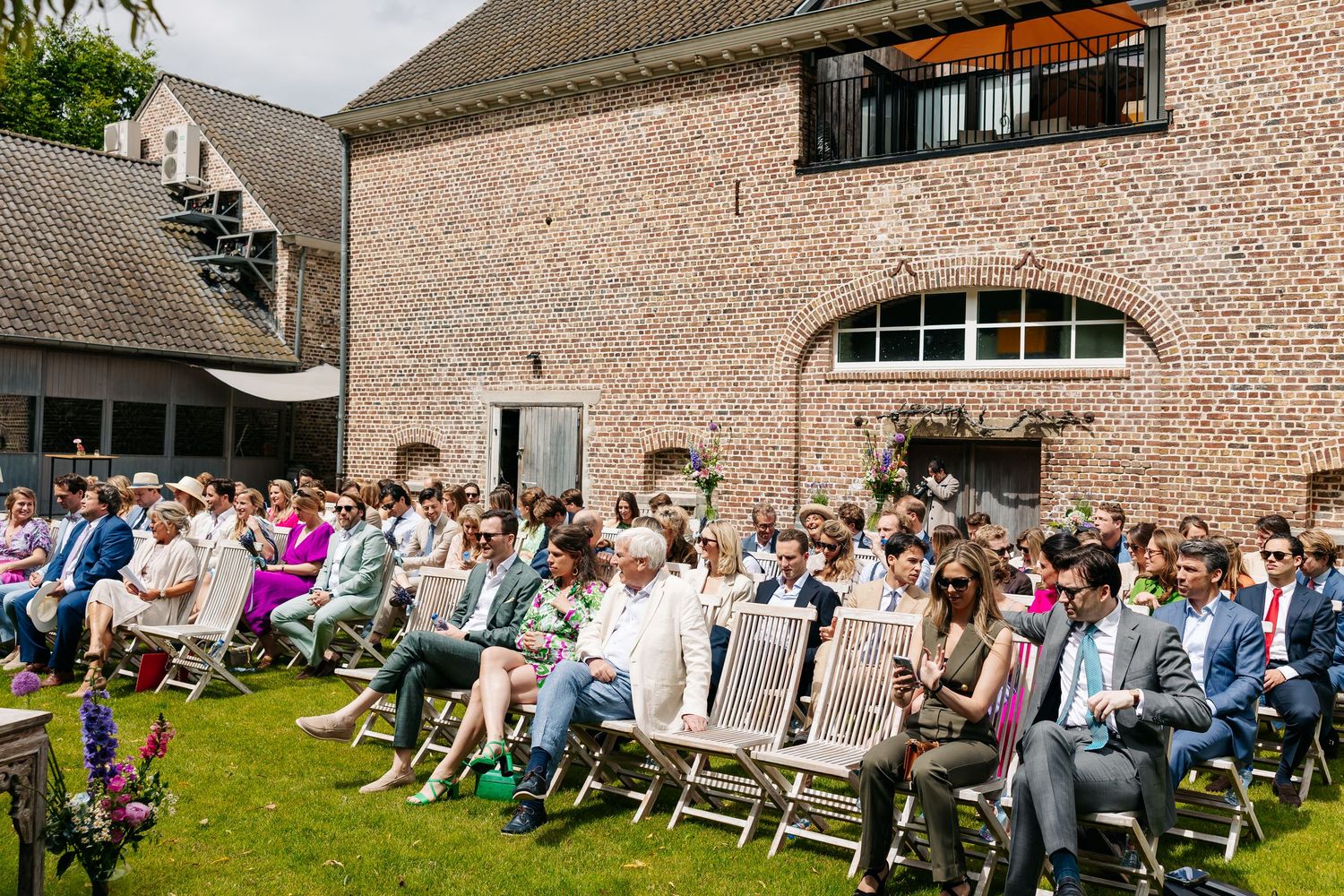 Wedding guests seated in white chairs outdoors during a sunny ceremony beside a rustic brick building.