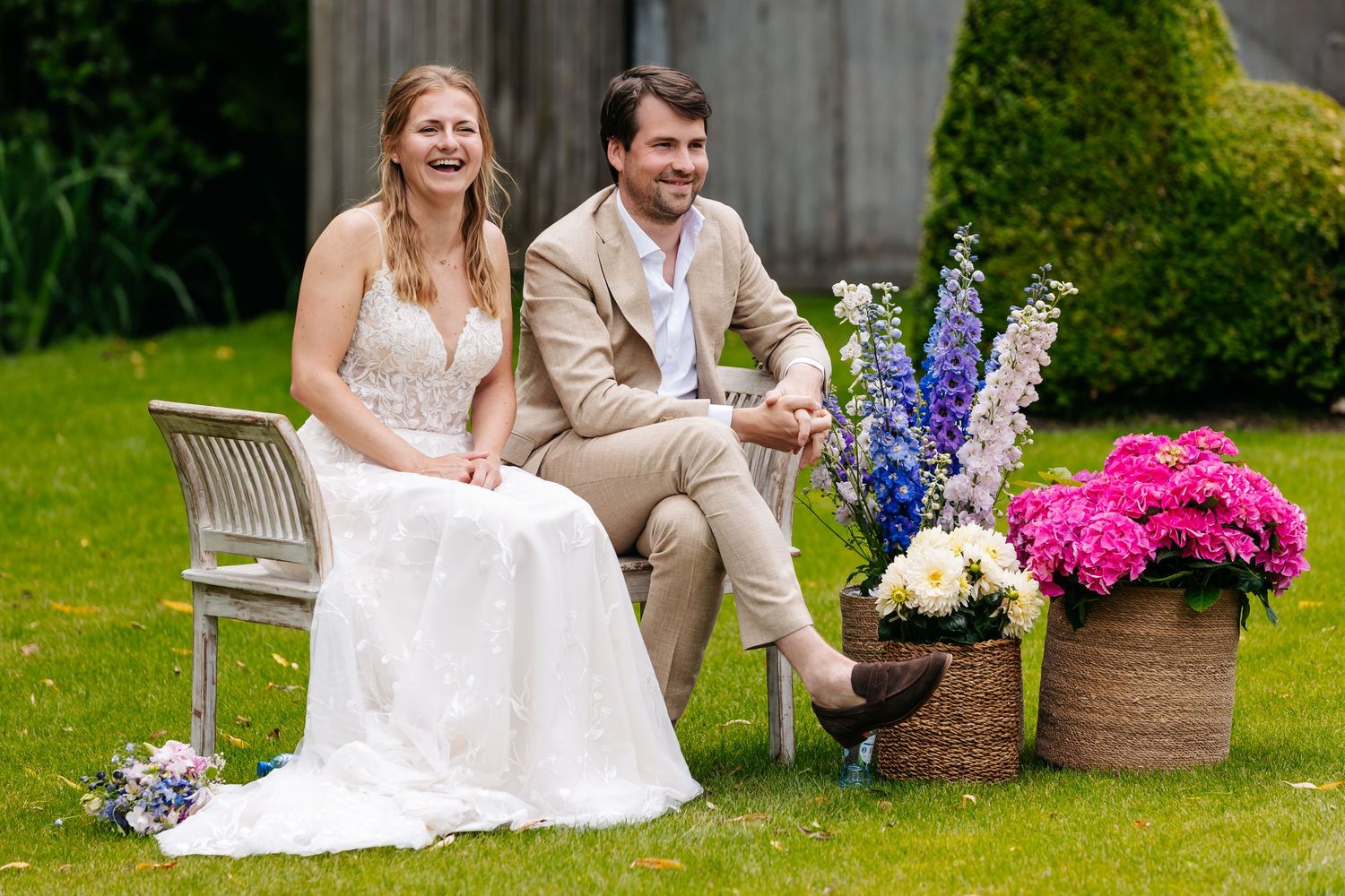 Bride in white gown and groom in beige suit laughing together on a bench surrounded by colorful flowers.