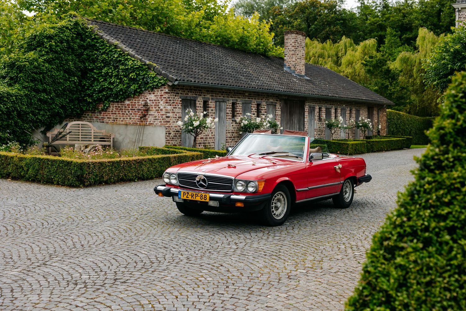 Red vintage Mercedes-Benz convertible parked on cobblestone driveway in front of a charming countryside estate.