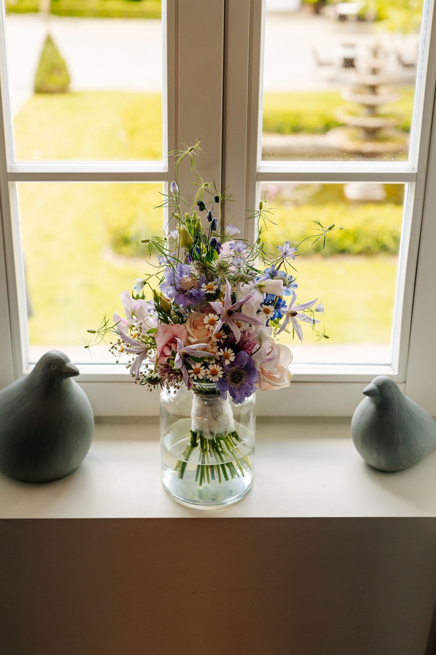 Wildflower wedding bouquet in glass jar on windowsill flanked by two ceramic bird figurines with garden view.