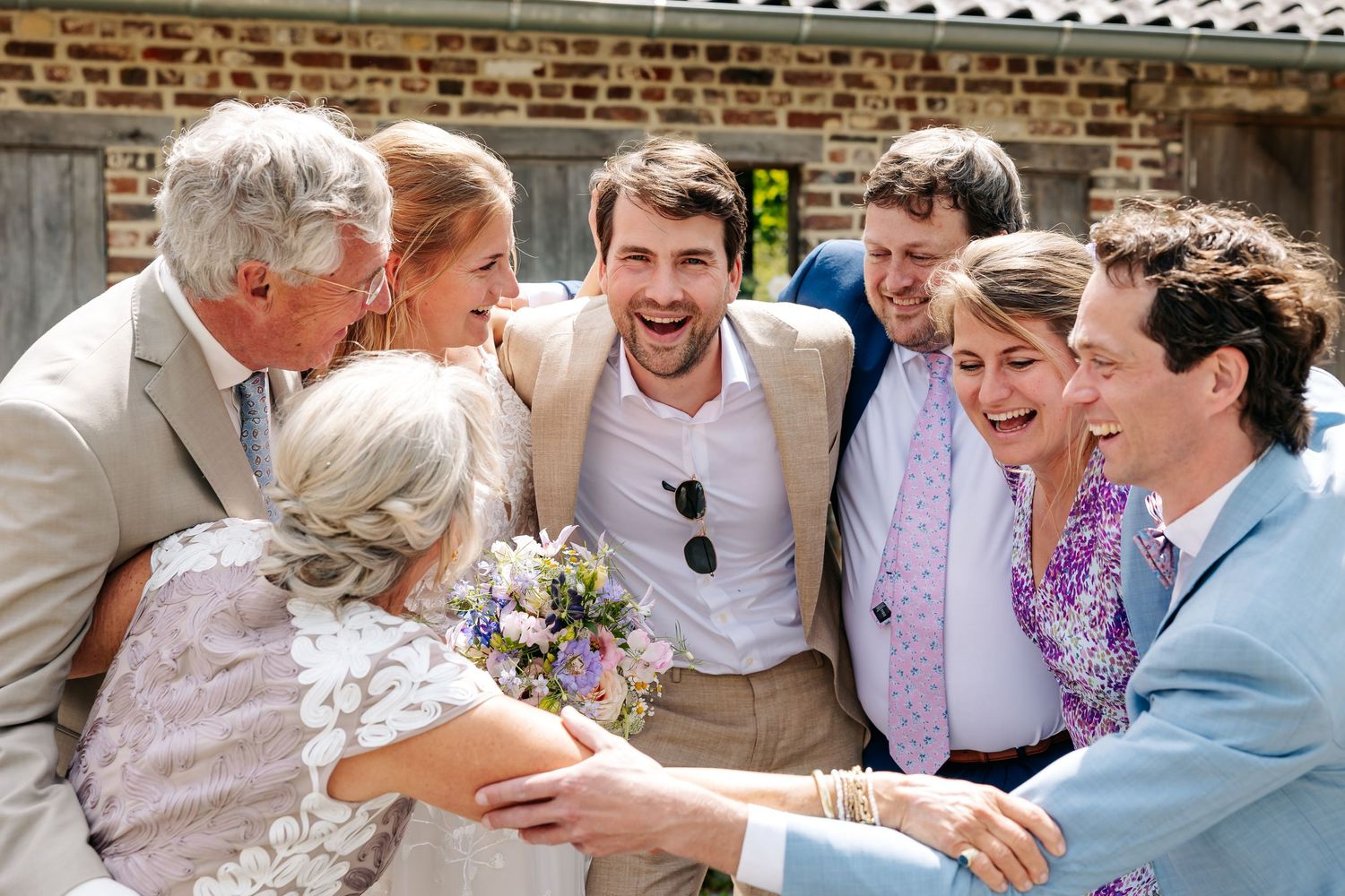 Joyful wedding guests gathering around laughing groom outdoors holding bridal bouquet.