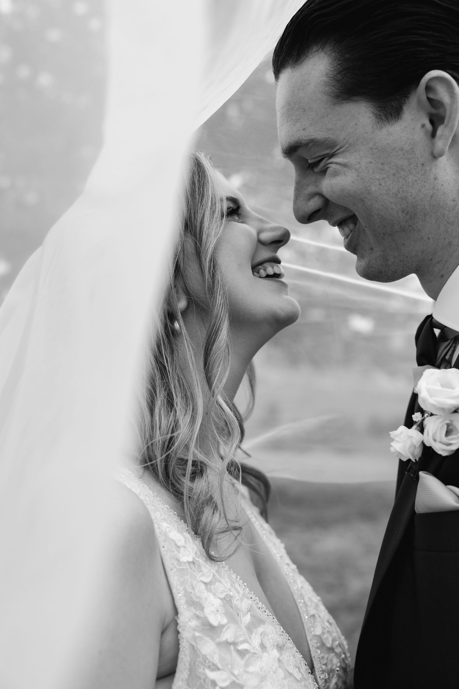 Black and white photo of smiling bride and groom gazing at each other under wedding veil.
