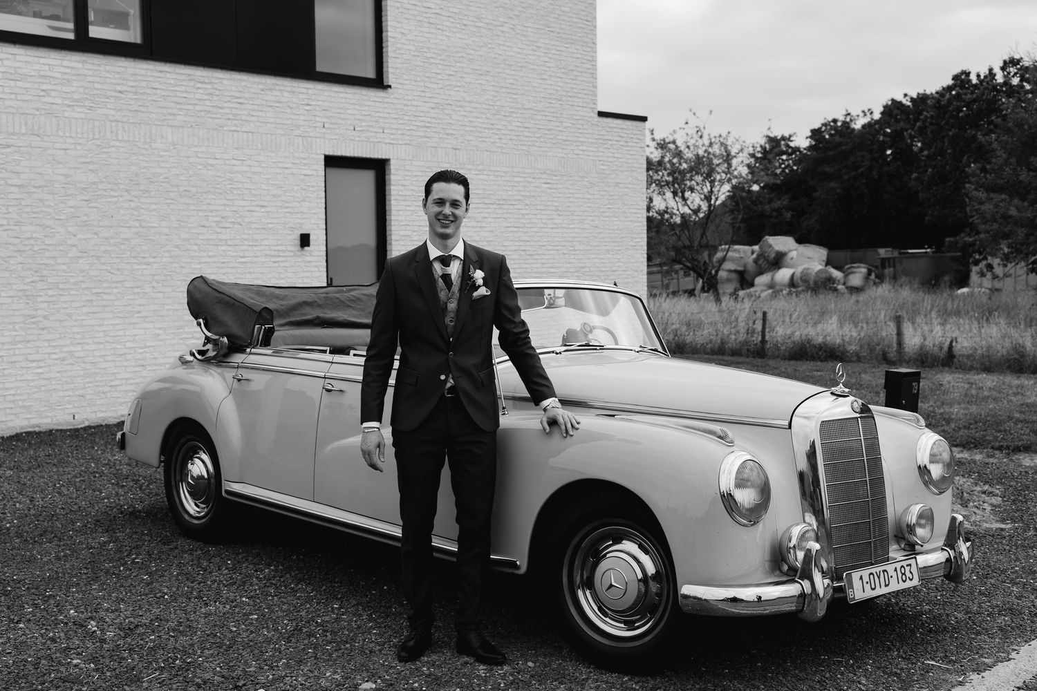 Groom in black wedding suit poses beside a classic vintage Mercedes convertible, black and white.