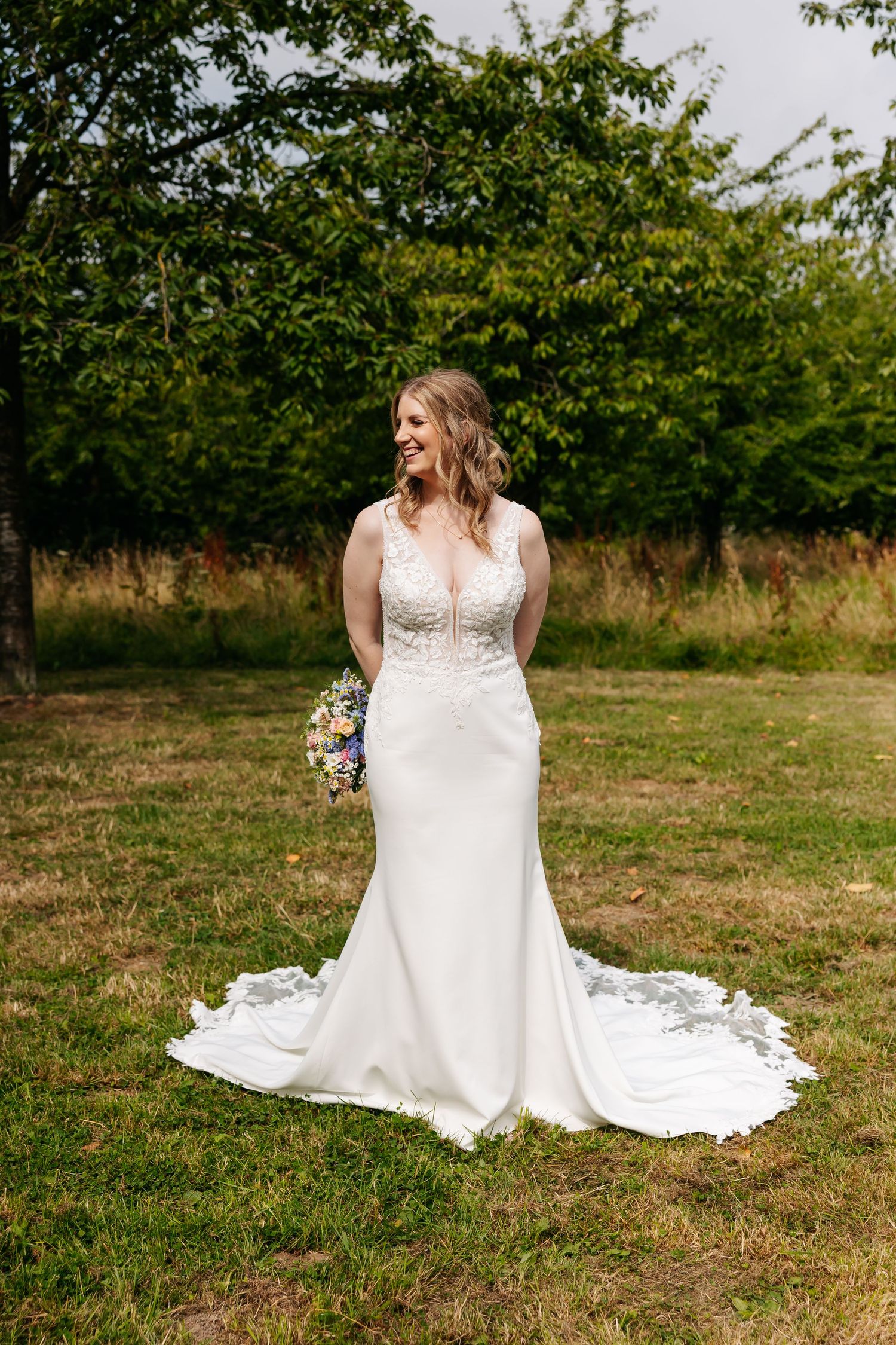 Bride in elegant lace wedding gown holding a colorful bouquet while standing on grass near green trees.