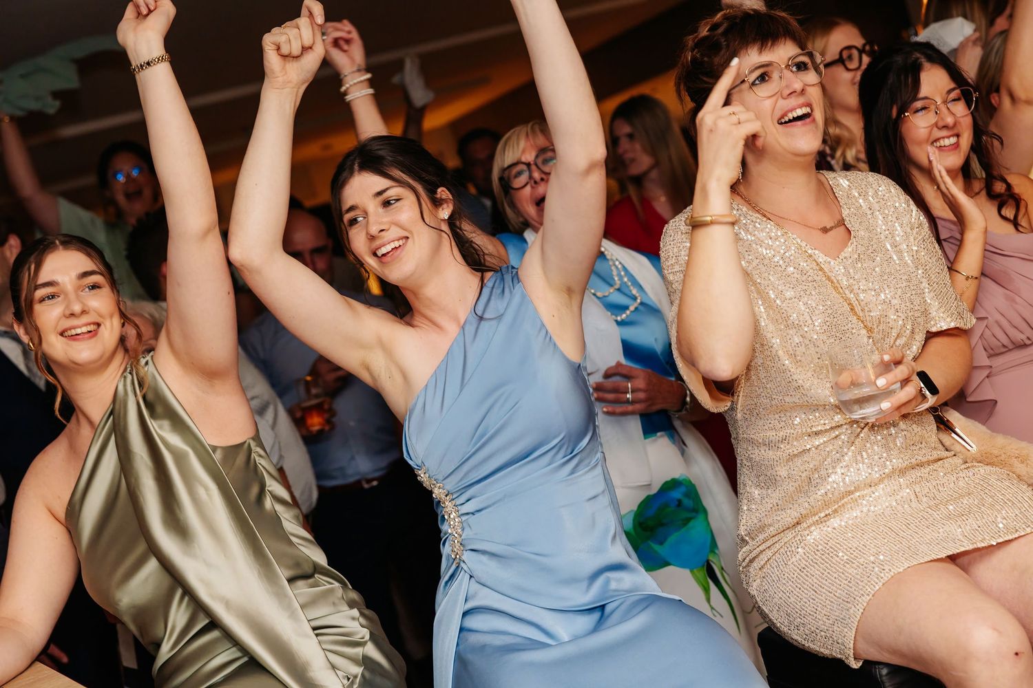 Wedding guests in colorful dresses celebrate joyfully on the dance floor at a reception.