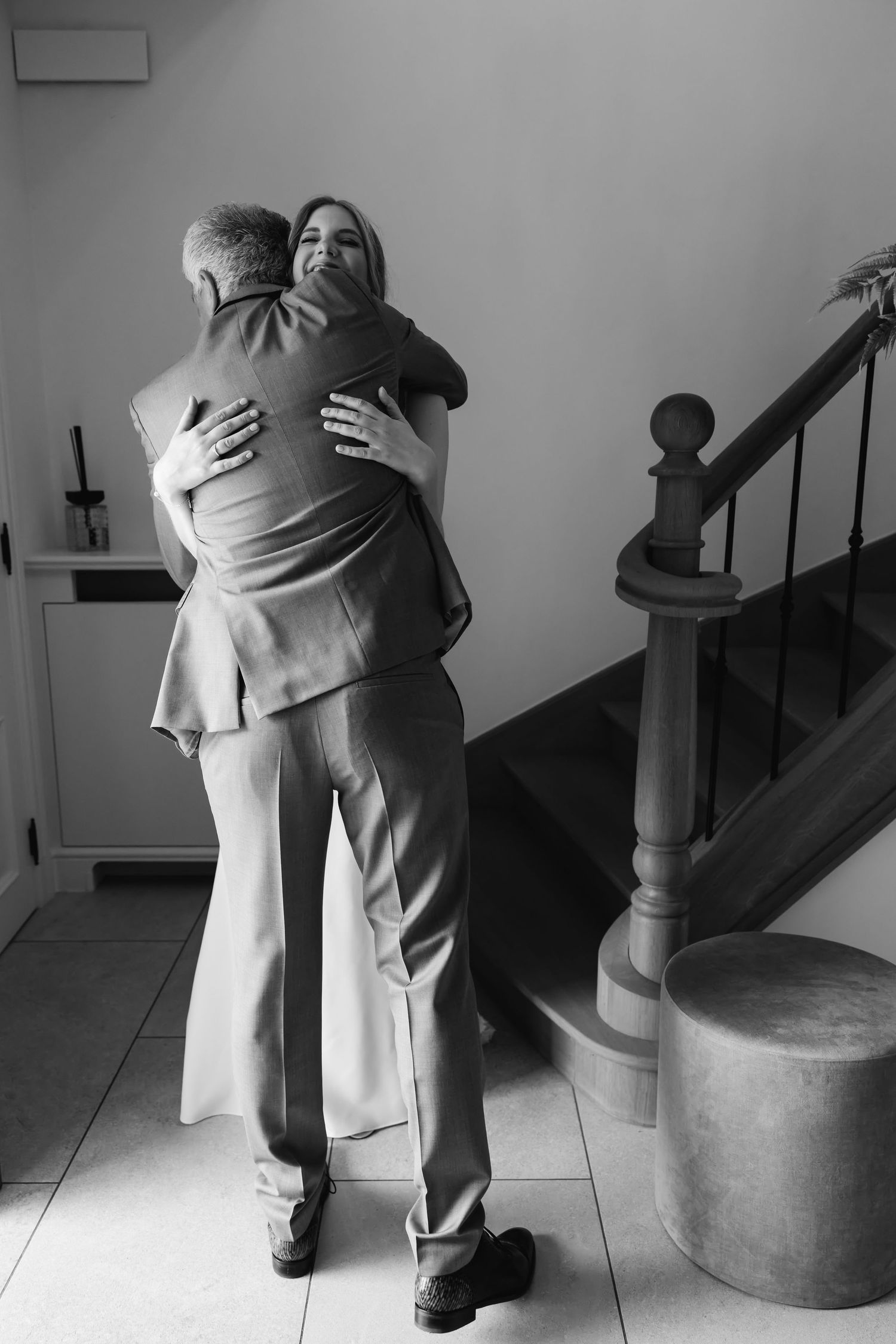 Black and white photo of bride and father sharing emotional hug near staircase on wedding day.
