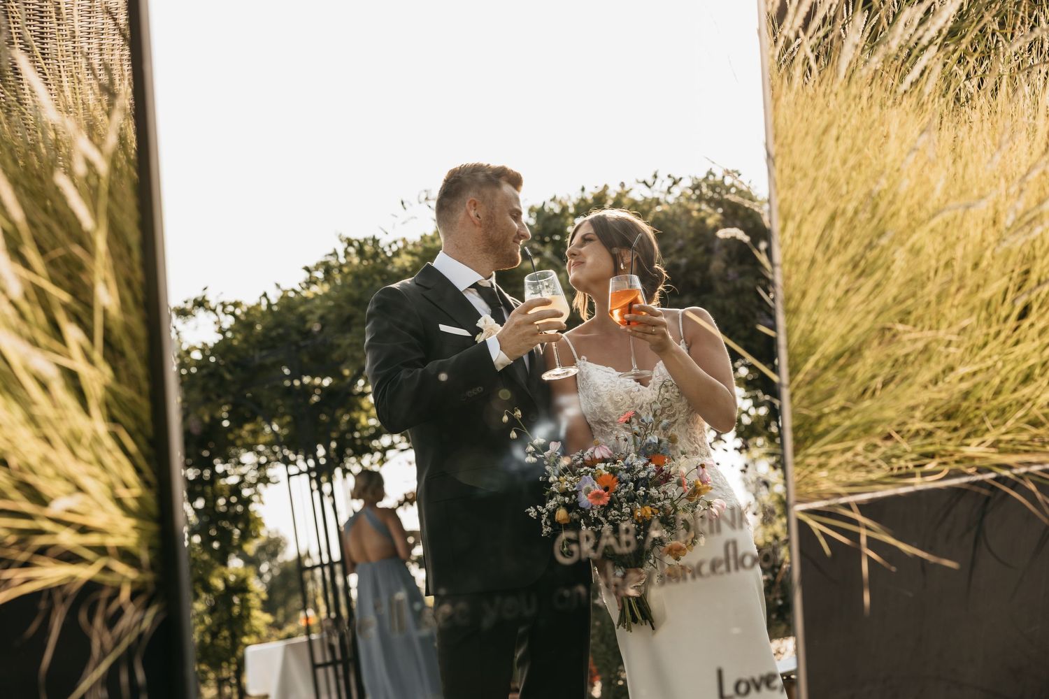 Bride and groom toasting with champagne glasses reflected in an outdoor mirror at their wedding reception.
