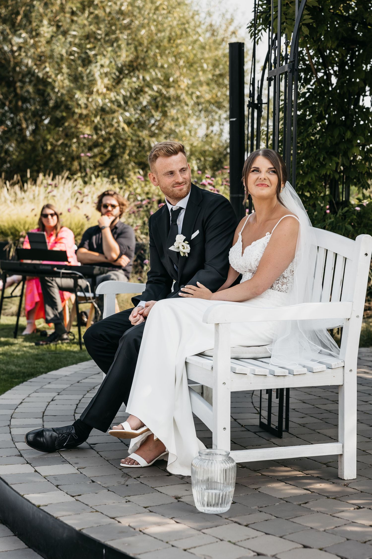 Bride in white gown and groom in black suit seated together on white bench during garden ceremony.