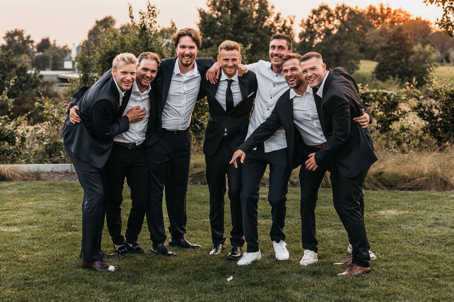 Groom and groomsmen in dark suits laughing together outdoors during wedding celebration.