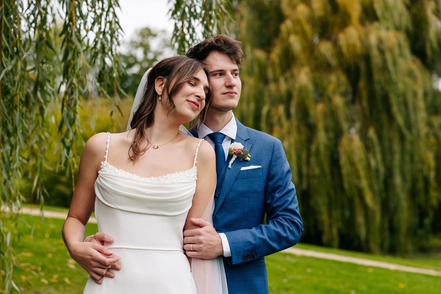 Bride in cowl neck white gown and groom in blue suit pose together under weeping willow tree.