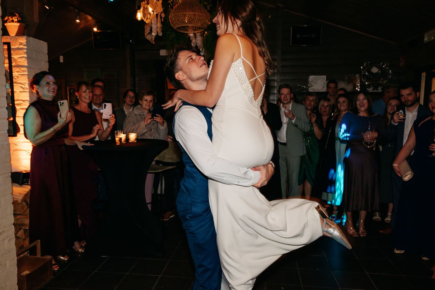 Groom lifts bride during first dance at wedding reception while guests cheer and capture the romantic moment.