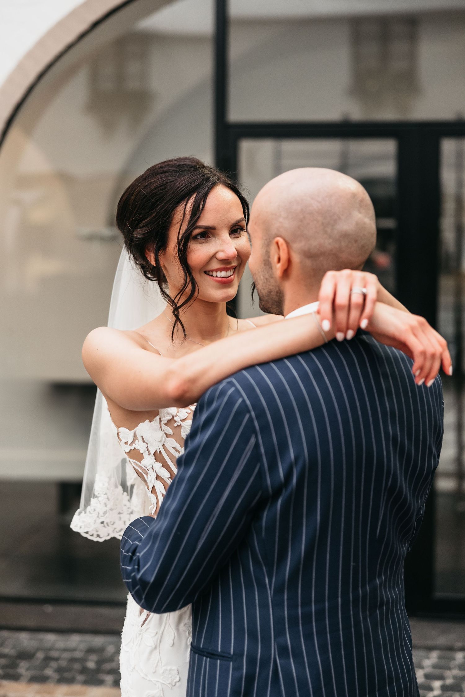 Radiant bride in white lace gown smiles at bald groom while embracing during wedding portraits.