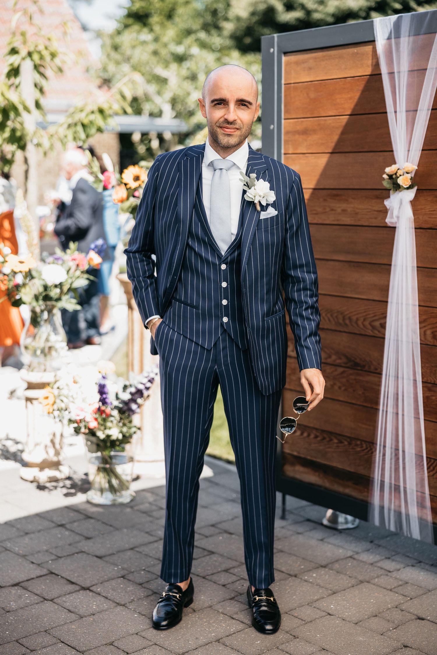 Bald groom smiling in navy pinstripe suit with white boutonniere at decorated outdoor wedding venue.