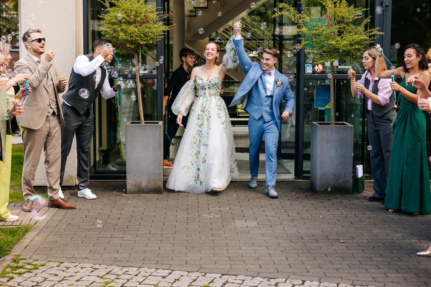 Joyful bride and groom in floral dress and blue suit celebrated by cheering guests outside venue.