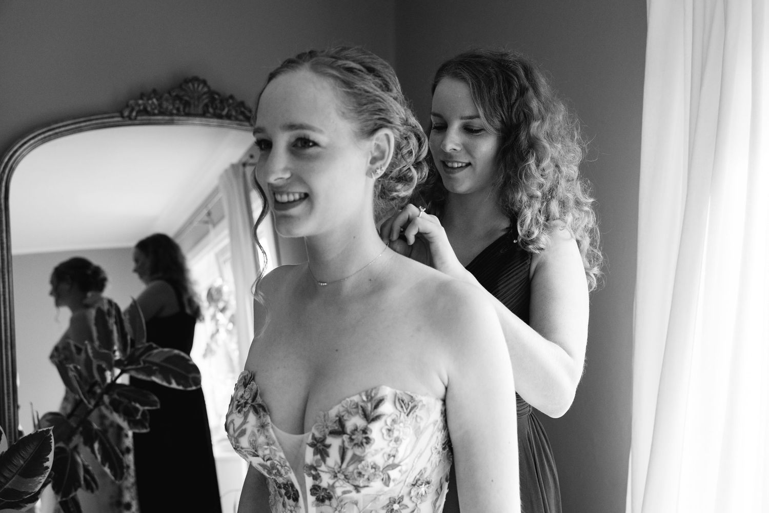 Black and white photo of bridesmaid helping smiling bride with earring before wedding ceremony.