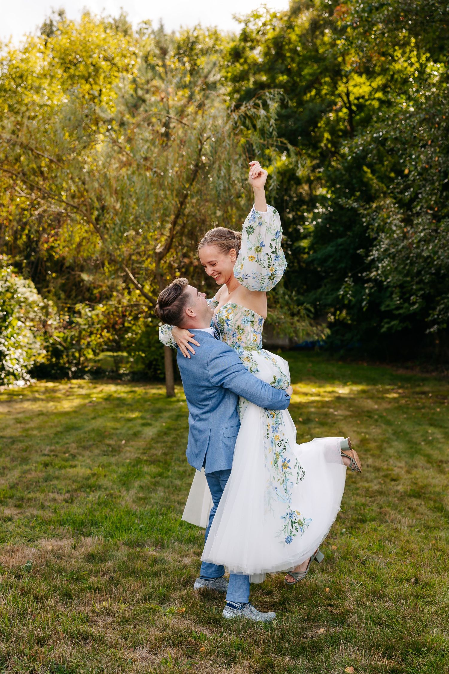 Groom in blue suit lifts laughing bride in floral wedding gown while dancing outdoors in green garden.