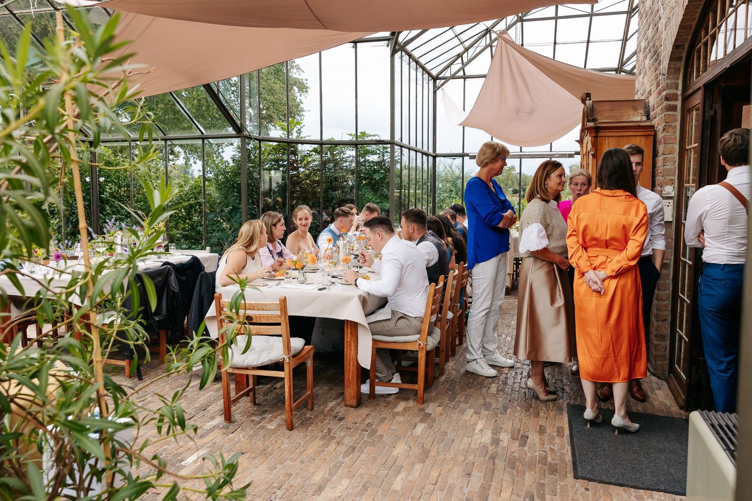 Wedding guests dining at decorated tables inside a bright sunlit glass conservatory venue.