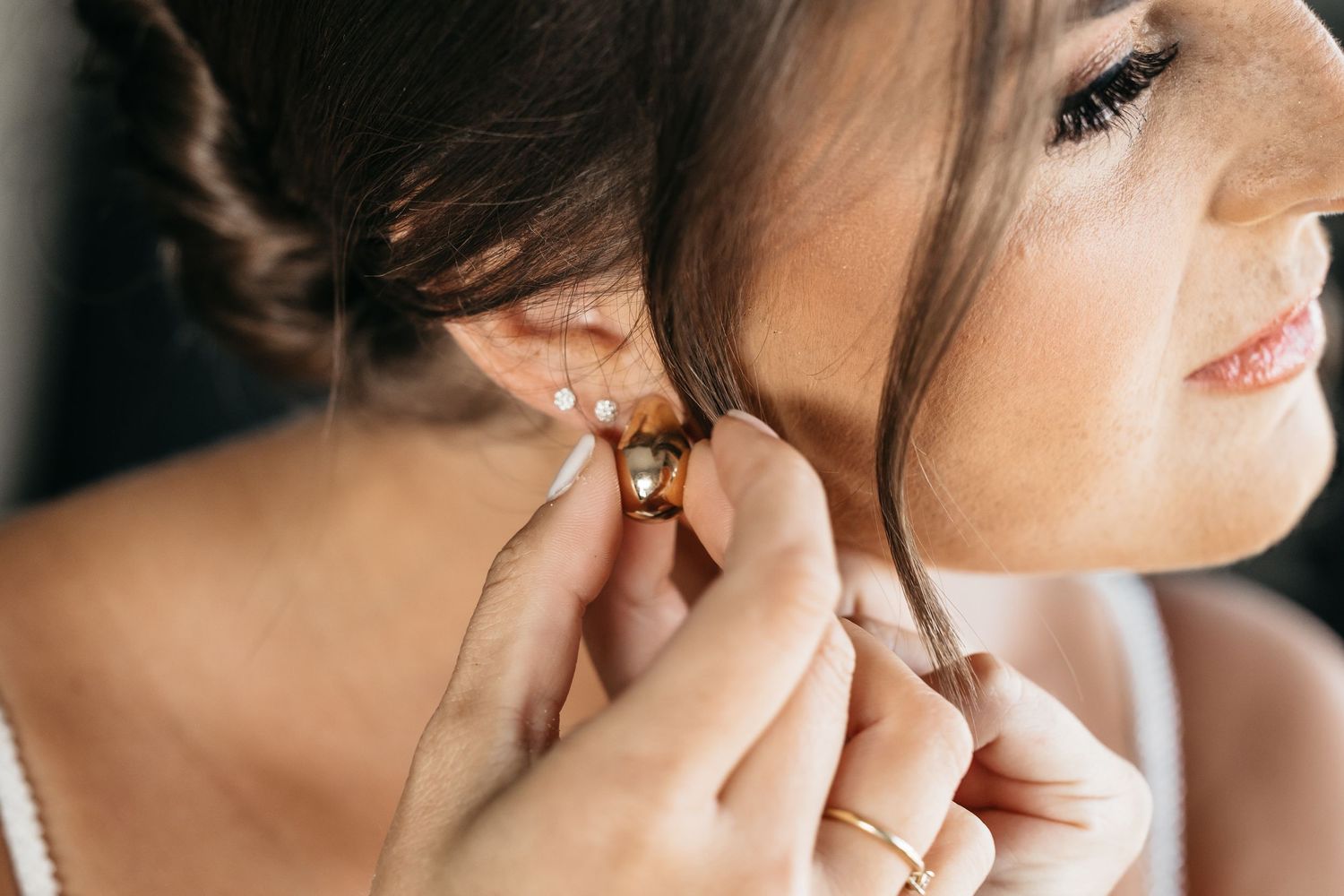 Close-up of hands placing gold earring on bride with elegant updo hairstyle.