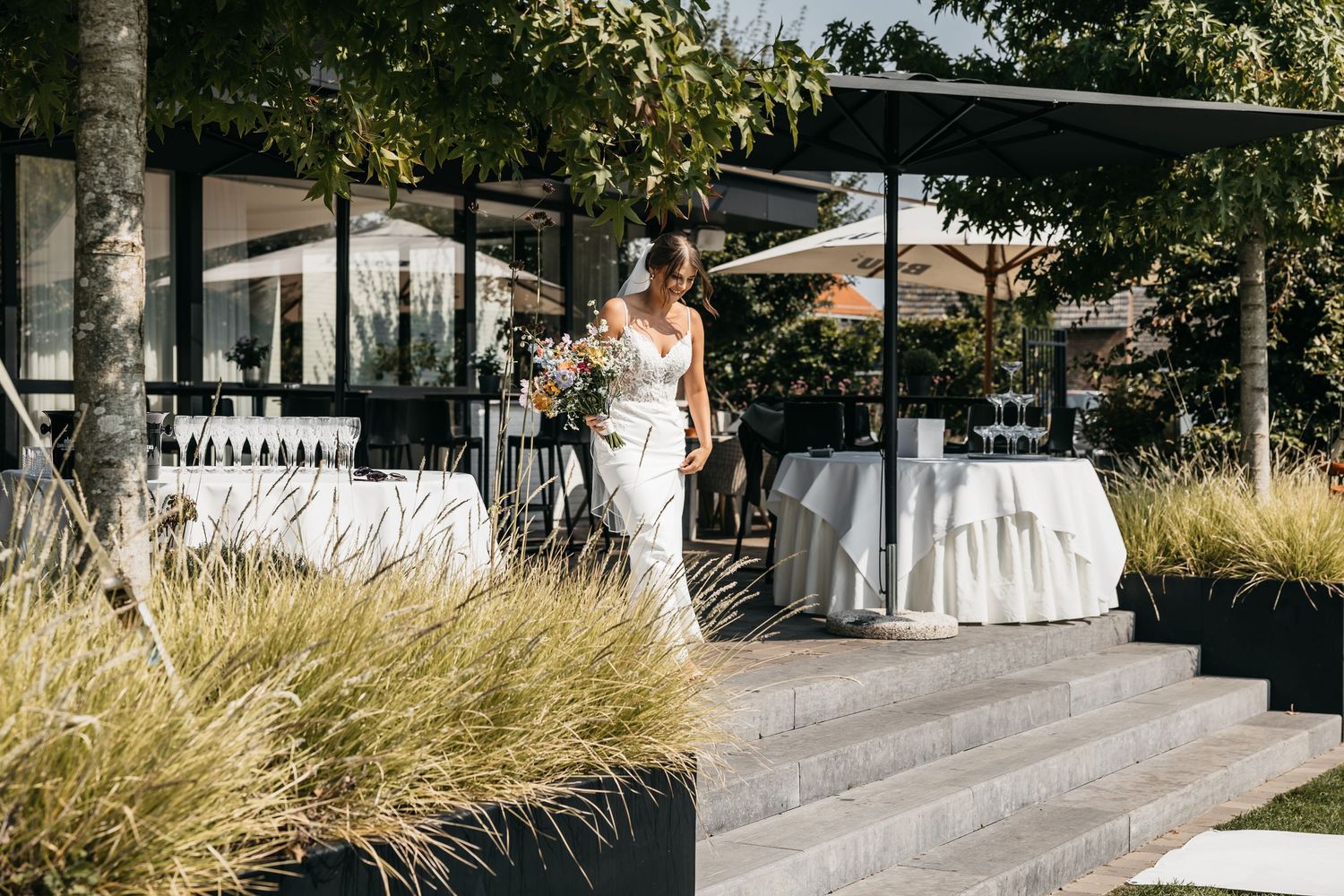 Elegant bride in white gown walks down steps carrying a colorful bouquet at a modern outdoor wedding venue.