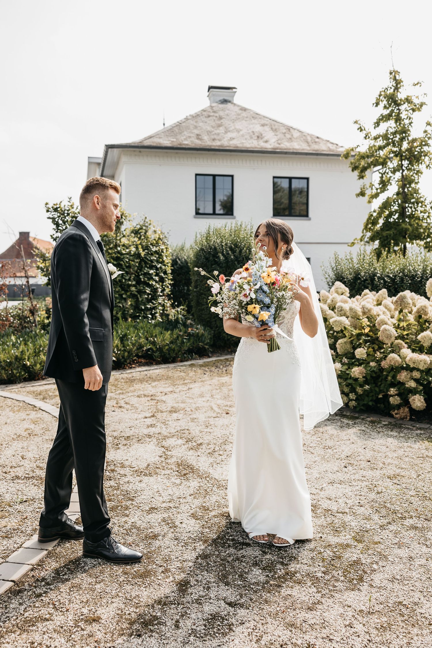 Bride smiles behind colorful wildflower bouquet while facing groom in garden setting.