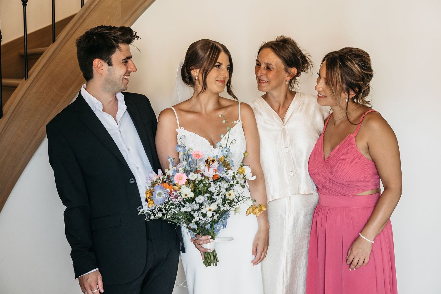 Bride holding colorful wildflower bouquet poses with three family members near wooden staircase.