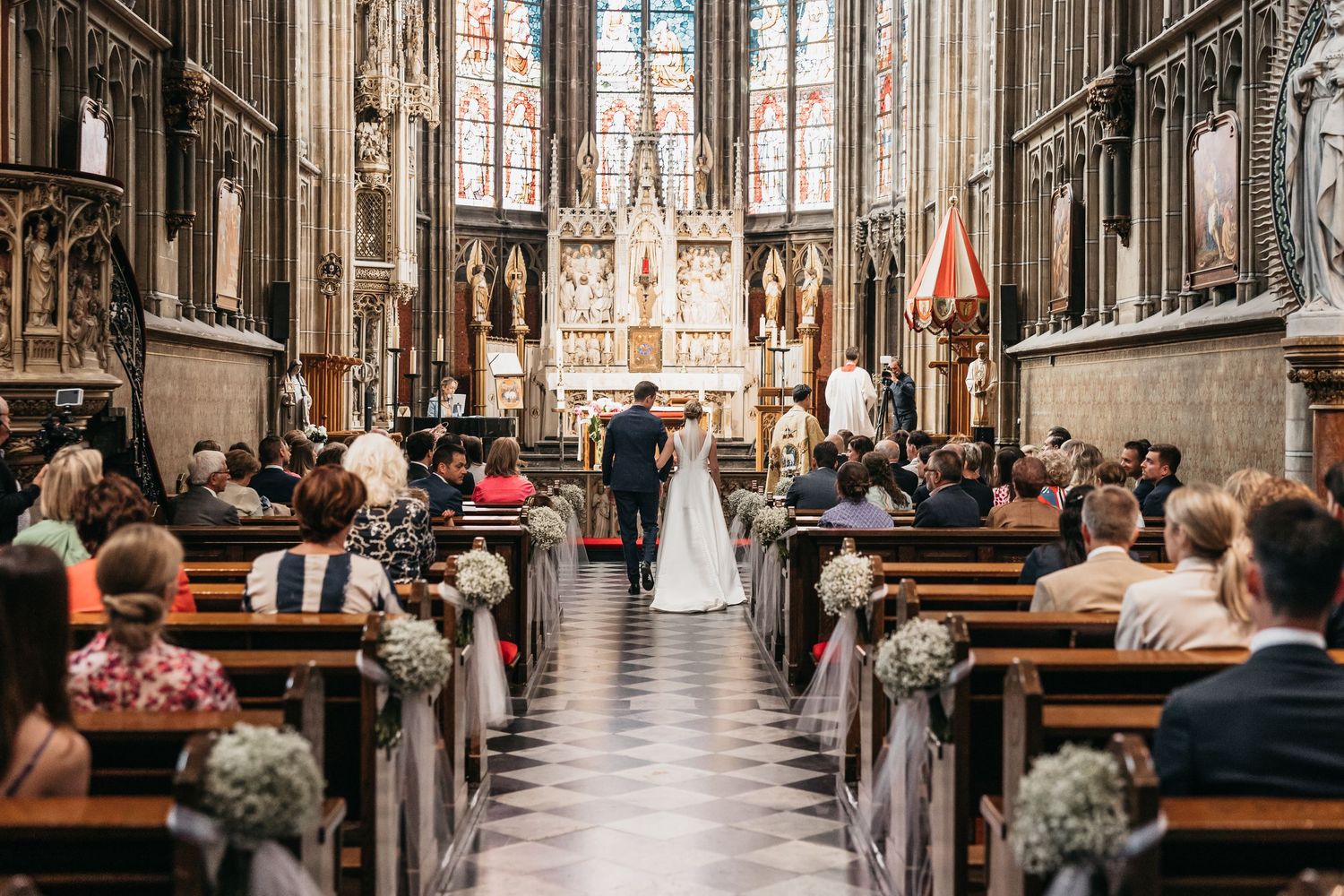 Wedding couple at altar of stunning Gothic cathedral with guests filling ornate wooden pews.