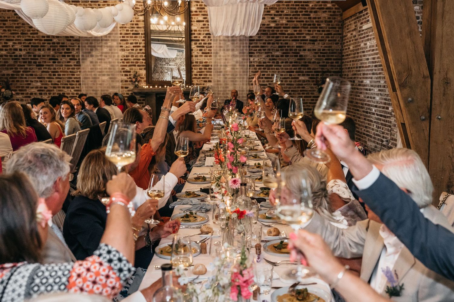 Wedding guests raising champagne glasses in a toast at a long reception table in a brick venue.