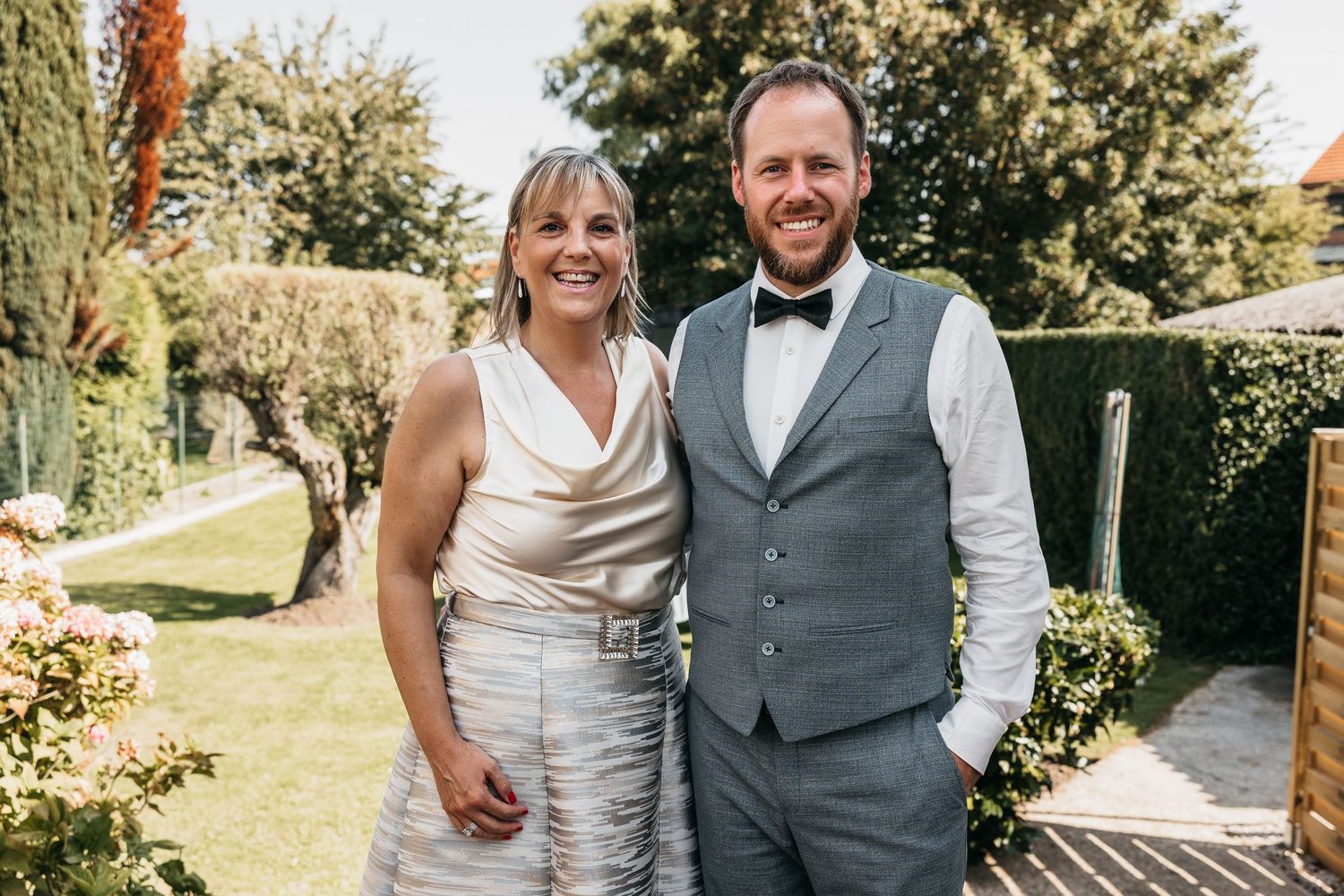 Smiling couple posing together outdoors at a wedding, man wearing gray vest and bow tie, woman in white dress.