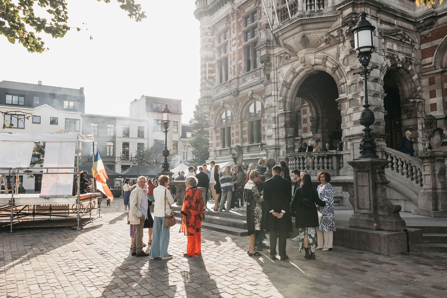 Wedding guests gather outside ornate historic building in golden sunlight on cobblestone plaza.