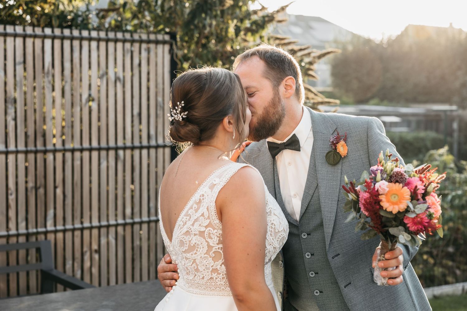 Bride and groom share a romantic kiss outdoors at golden hour, groom holding vibrant pink and orange bouquet.
