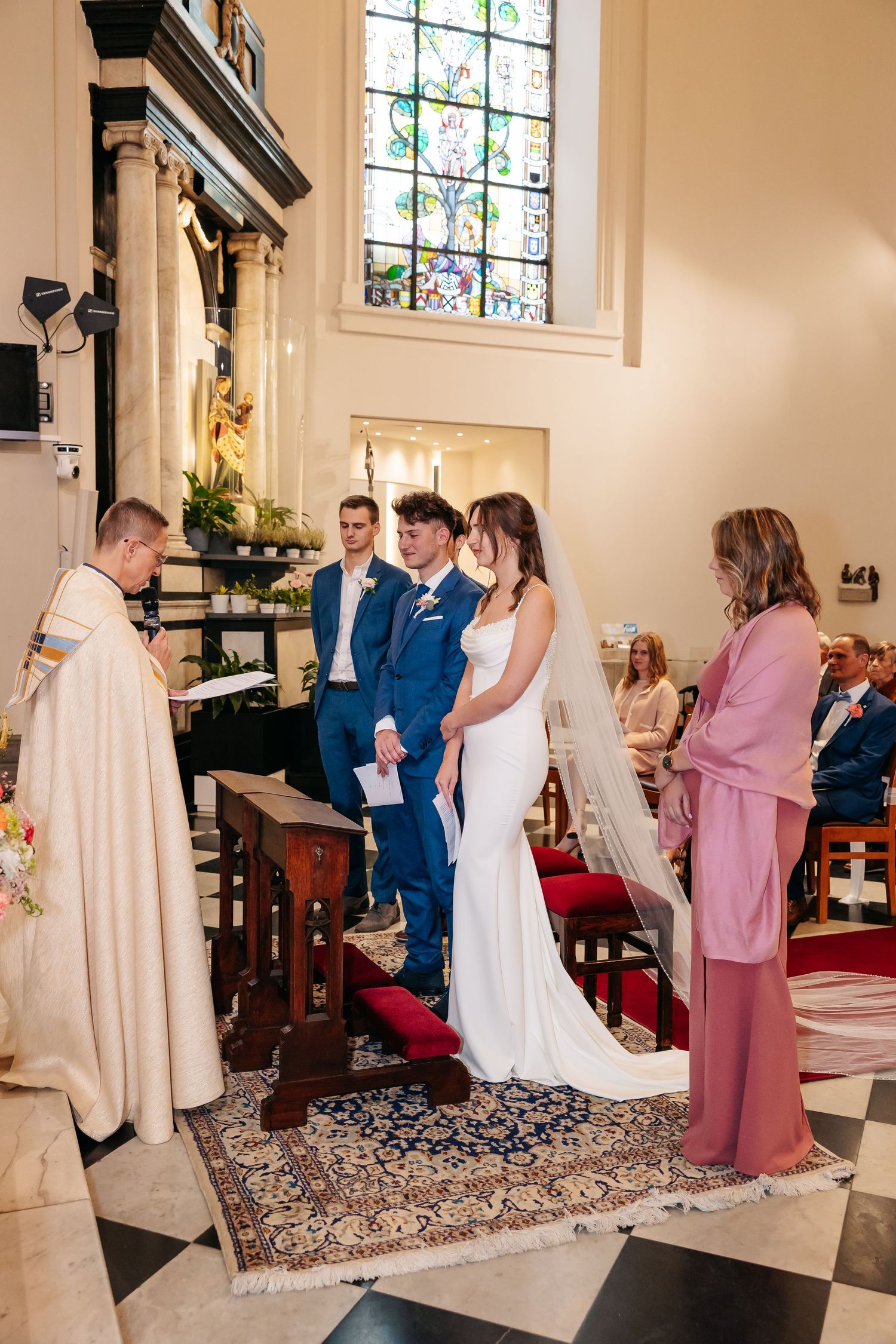Bride and groom exchange vows before officiant on ornate rug in sunlit church ceremony.