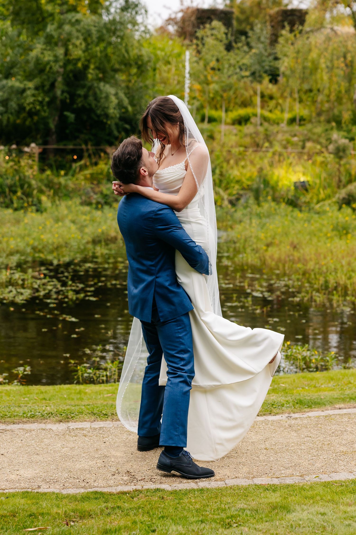 Groom sweeping bride off her feet in a romantic embrace by a lush garden pond.