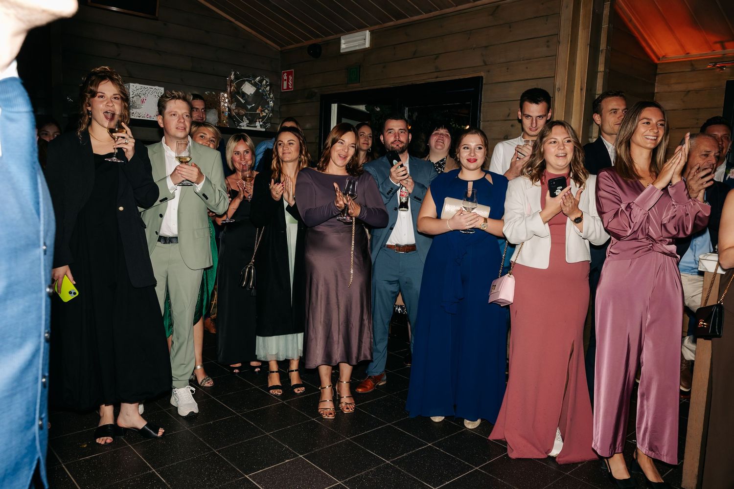 Wedding guests clapping and celebrating during reception, dressed in elegant formal attire in a warmly lit venue.