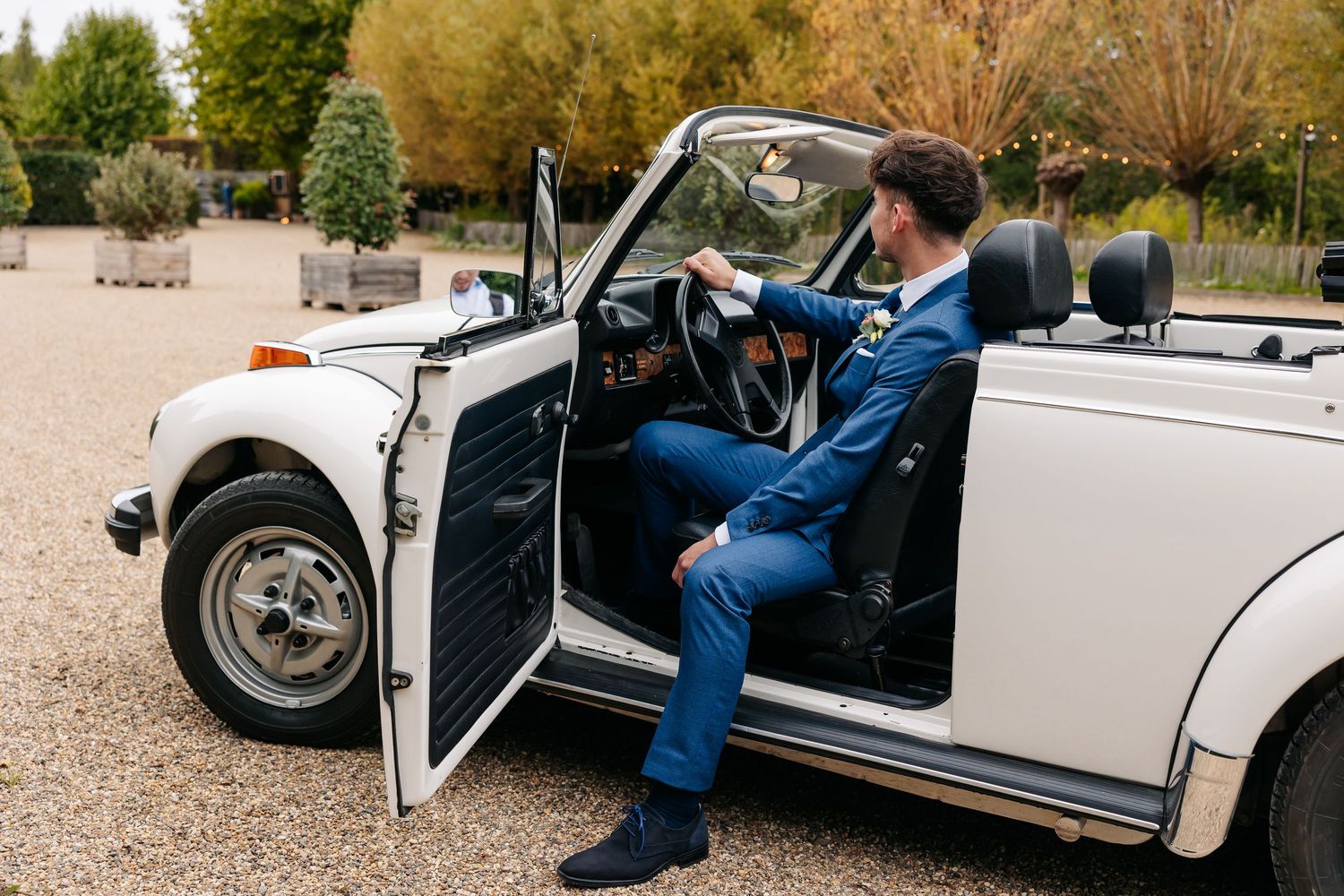 Groom in blue suit sitting in open door of vintage white convertible car on gravel path.