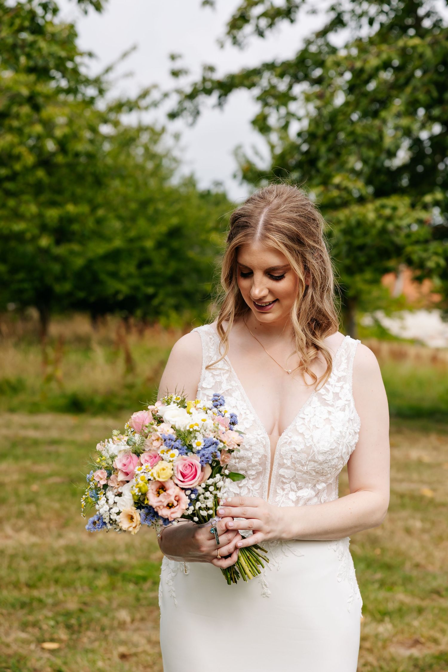 Bride in a deep V-neck lace gown gazing at a pastel floral bouquet in a garden.