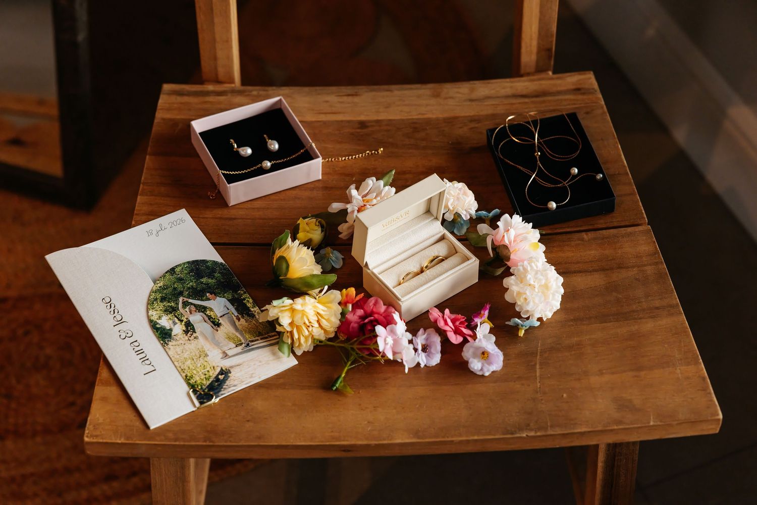 Wedding flat lay with rings, earrings, necklace, floral accessories and invitation on wooden table.