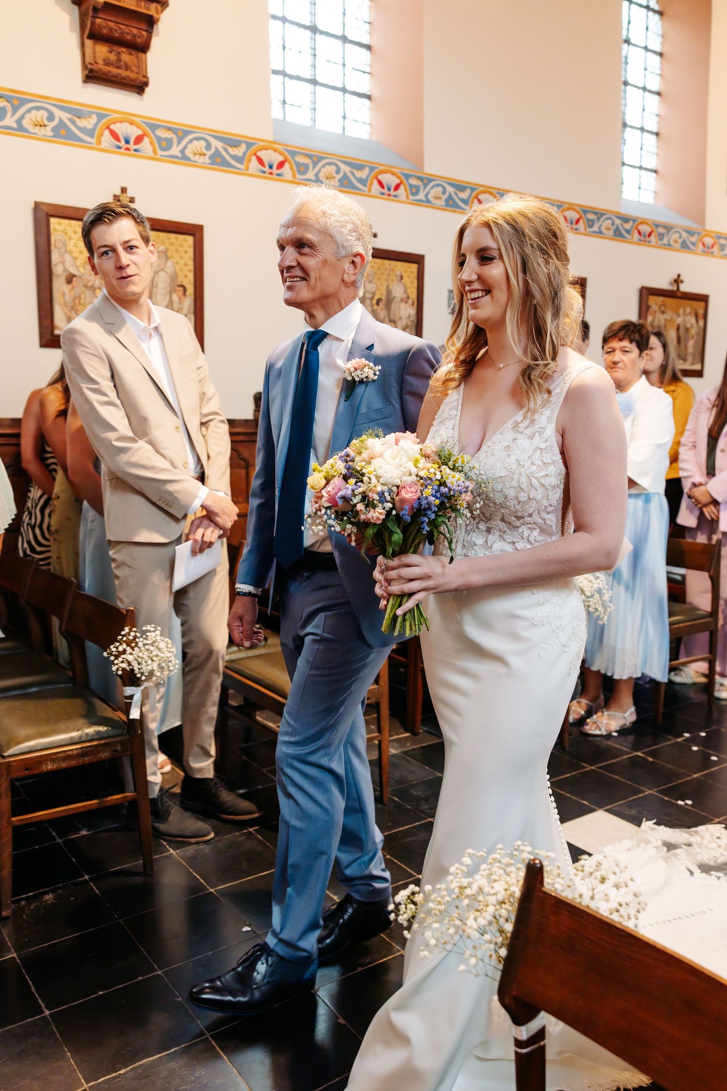 Bride walks down church aisle with escort holding colorful bouquet at wedding ceremony.