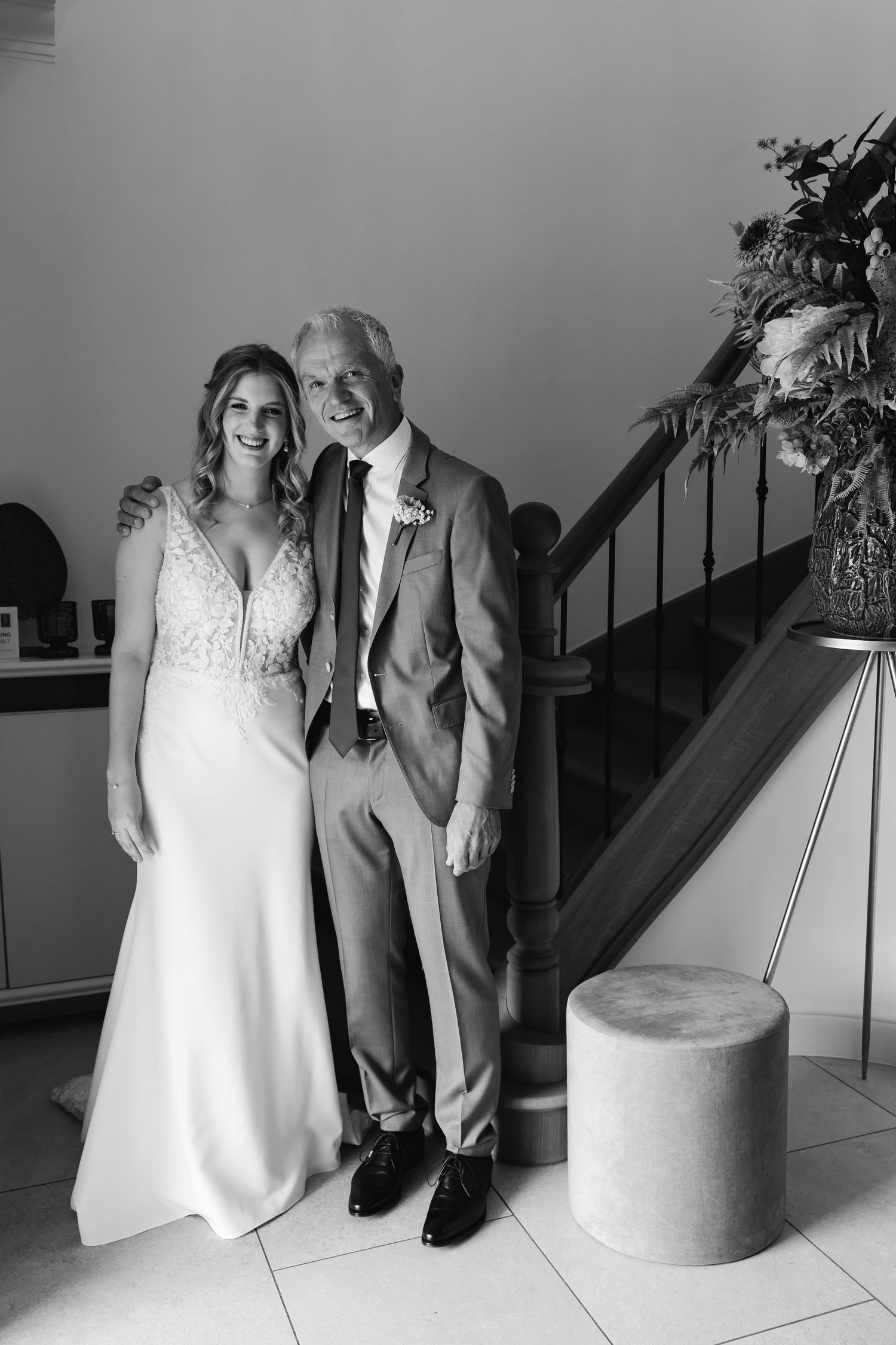 Black and white portrait of bride and older man smiling together beside staircase on wedding day.