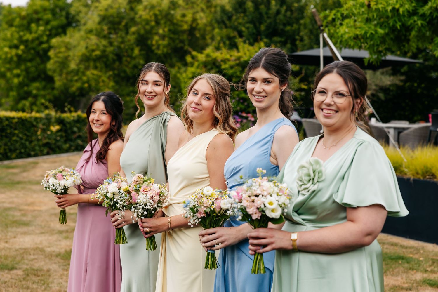 Five bridesmaids smiling in mismatched pastel gowns holding bouquets at sunny outdoor wedding.